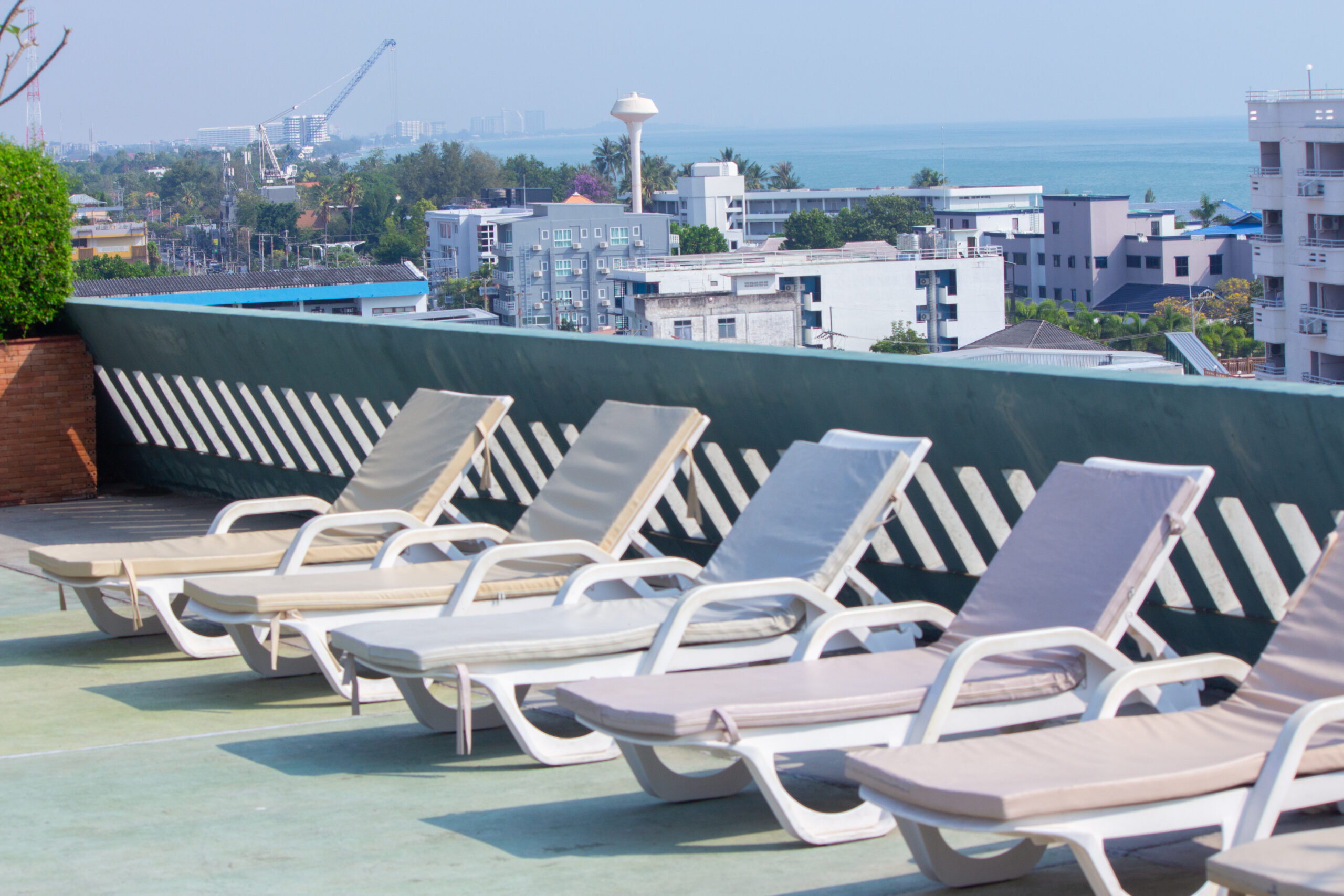A row of white sun loungers with beige cushions on a rooftop sundeck overlooking the city and ocean at Hisea Hua Hin Hotel.